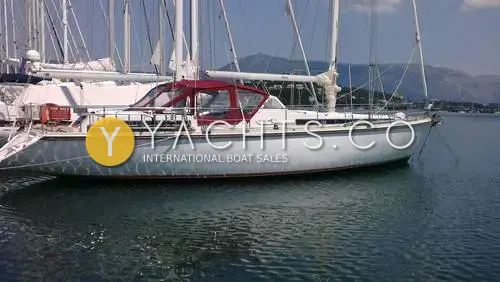Amel Super Maramu 2000: starboard side of the white ketch moored in calm water with a red cockpit canopy and mountains behind