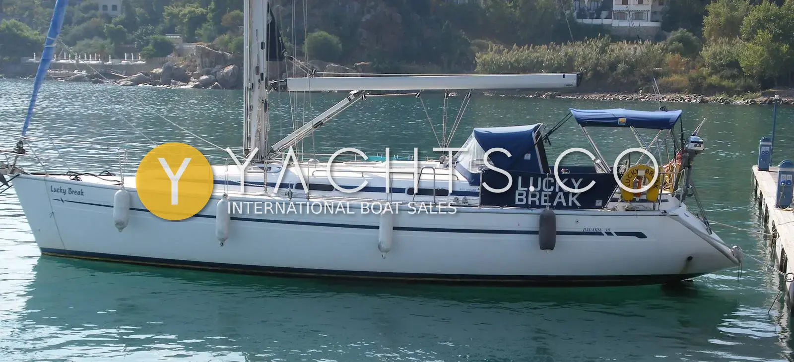 Bavaria 38: white hull afloat at a quay with blue sprayhood, wooded shoreline behind