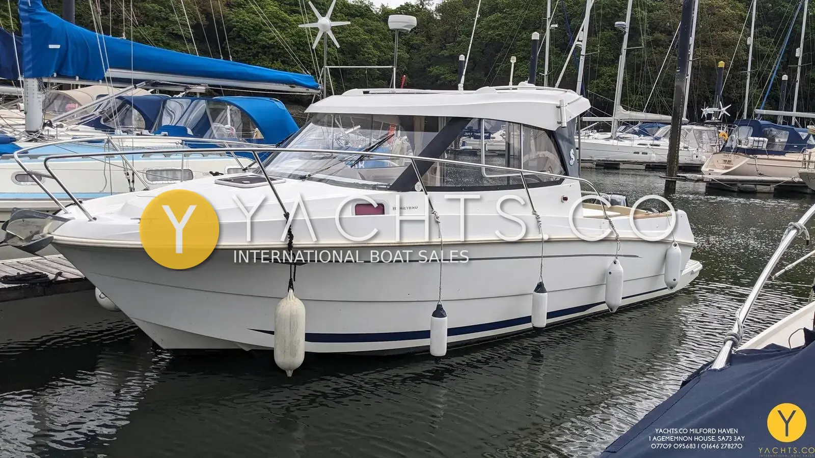 Beneteau Antares 7.80: white hull and wheelhouse seen from the port bow, moored in a wooded marina