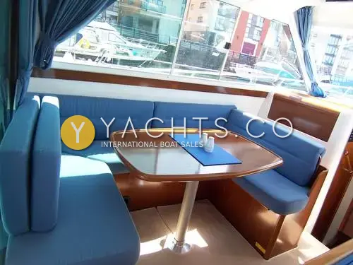 Beneteau Antares 9: saloon U-shaped dinette in blue upholstery with varnished table and marina view through windows