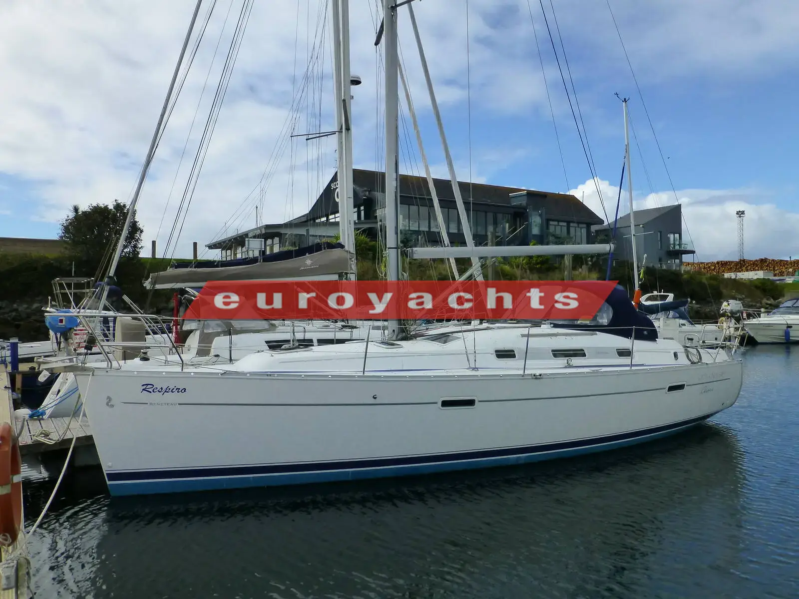 Beneteau Oceanis 343: white hull with blue boot stripe and blue sprayhood, berthed in a marina beside a modern building