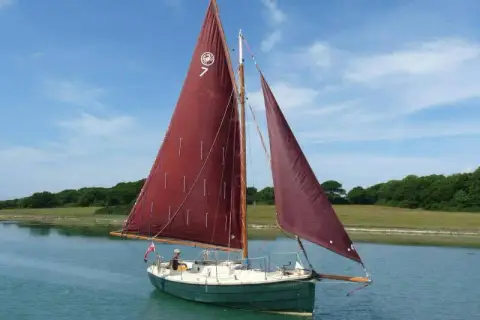 Cornish Crabber 22 Gaff Cutter: under sail with dark tan gaff main and headsails on a calm tree-lined estuary