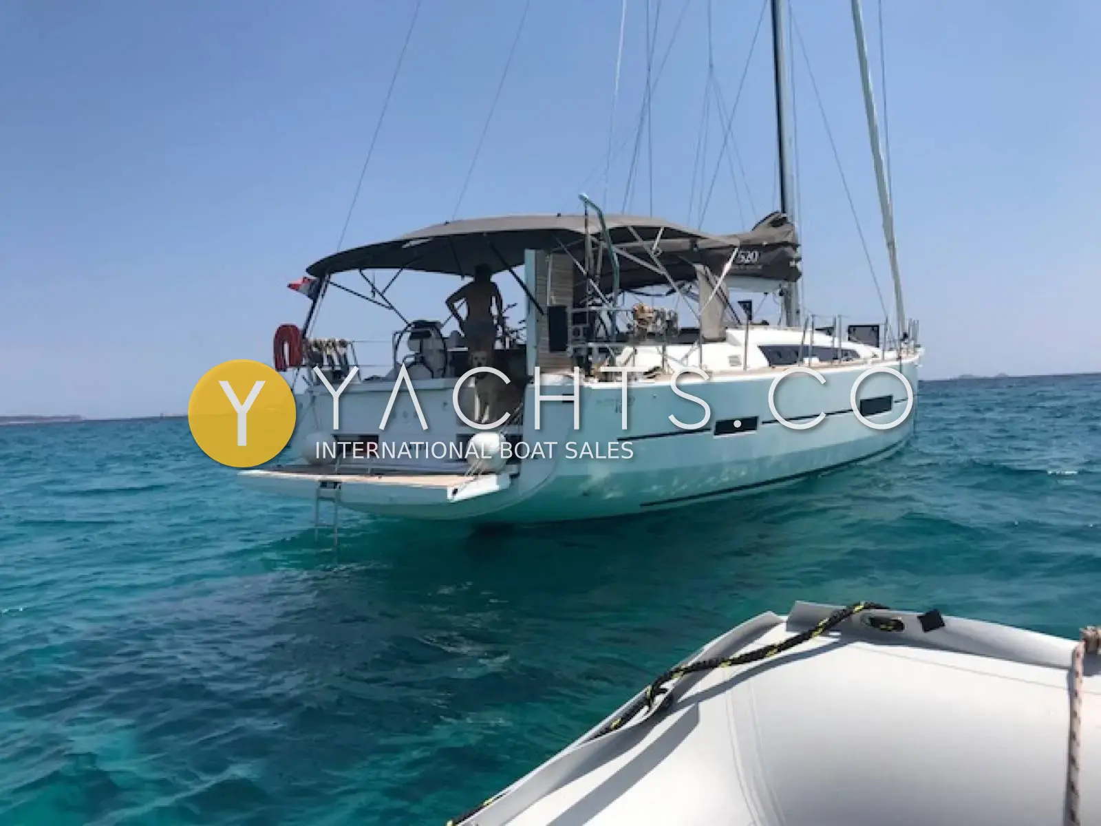 Dufour 520 Grand Large: stern view at anchor in turquoise water, bimini up and swim platform lowered, seen from tender