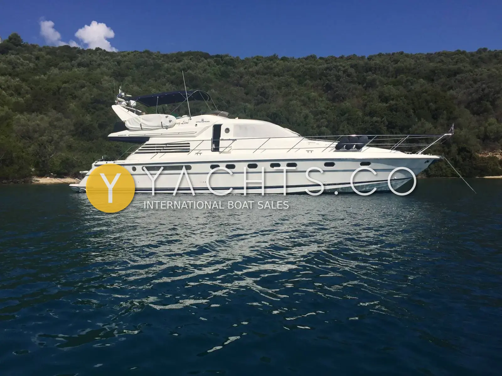 Fairline Squadron 62/65: starboard profile at anchor in a calm bay below wooded hills, flybridge bimini up