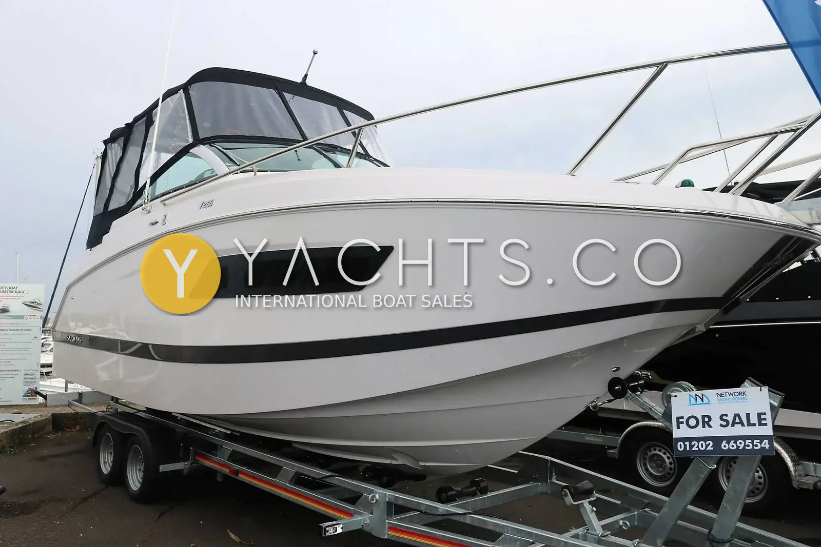 Four Winns Vista 255: starboard bow view of the white hull on a twin axle trailer, black canopy fitted, ashore
