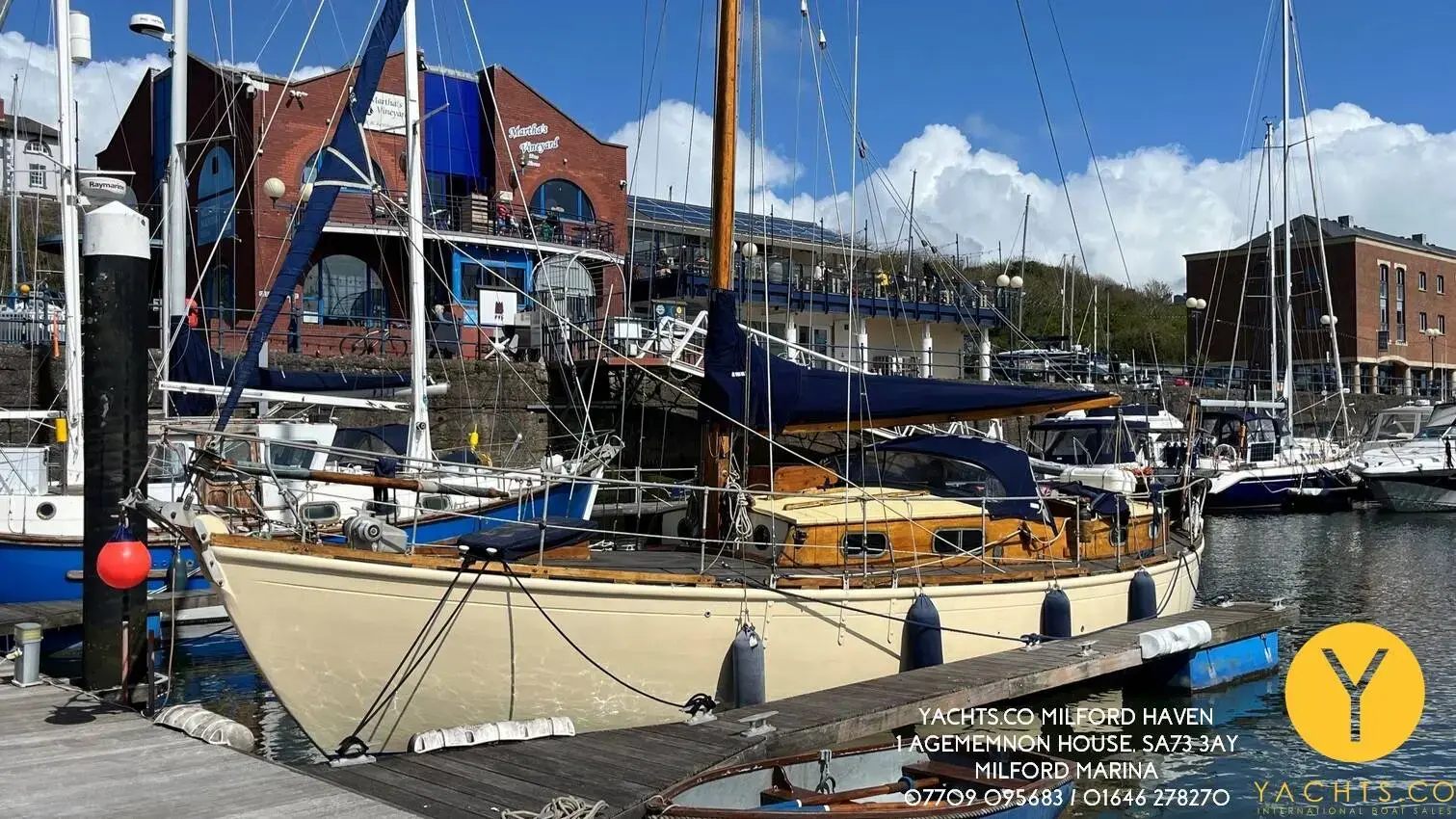 Hillyard 12 Ton: varnished wooden yacht with cream hull moored at a marina pontoon beside brick buildings