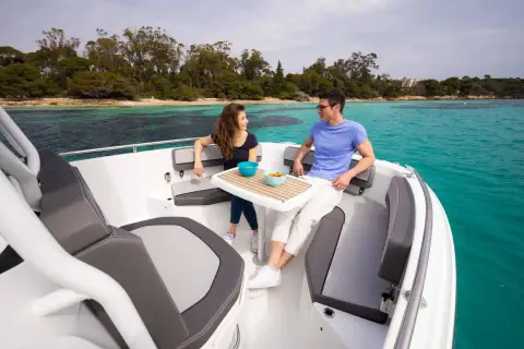 Jeanneau Cap Camarat 7.5 CC: forward cockpit seating with table set for drinks, two people relaxing near a wooded shore