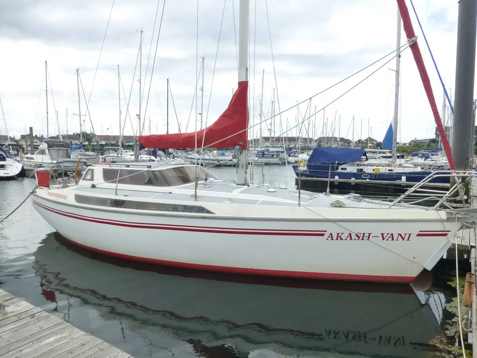 Jeanneau Espace 1000: white hull with red stripes and wheelhouse, moored in a marina with red sail cover