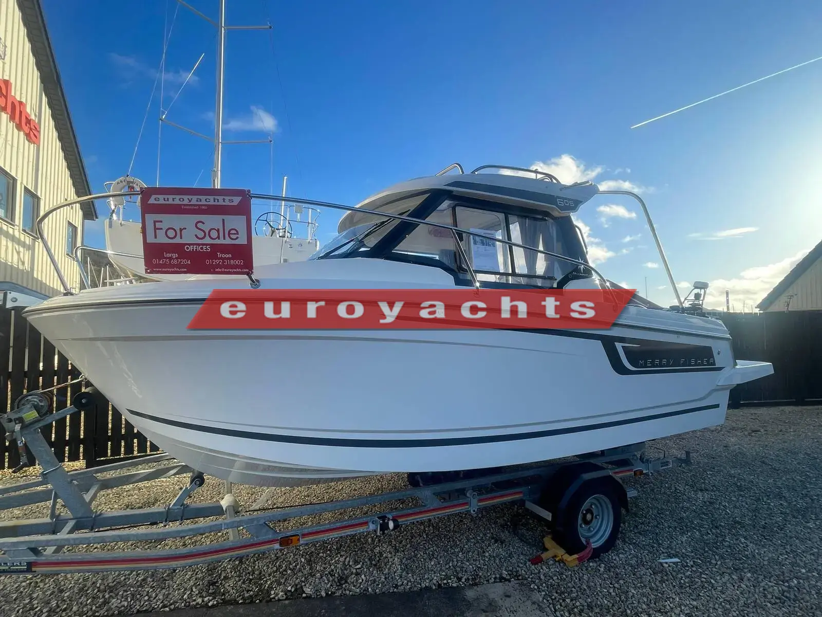 Jeanneau Merry Fisher 605: white hull with grey stripe on road trailer, ashore beside a dealership building under blue sky