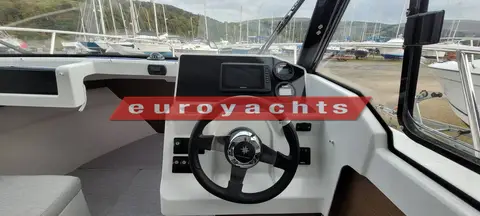 Jeanneau Merry Fisher 605: helm station with black steering wheel, dashboard console and view over boatyard through windscreen