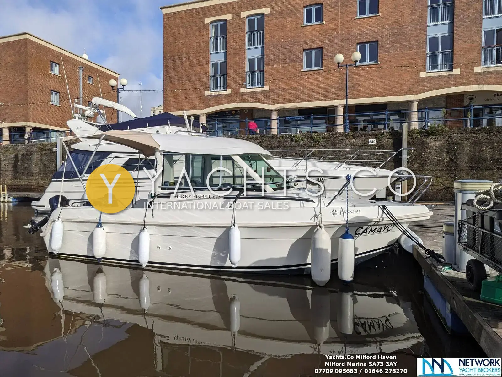 Jeanneau Merry Fisher 625: white hull afloat in a marina berth beside a red-brick waterfront building