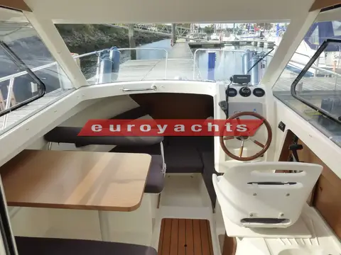 Jeanneau Merry Fisher 645: wheelhouse interior with dining table to port and wooden-rimmed wheel at starboard helm