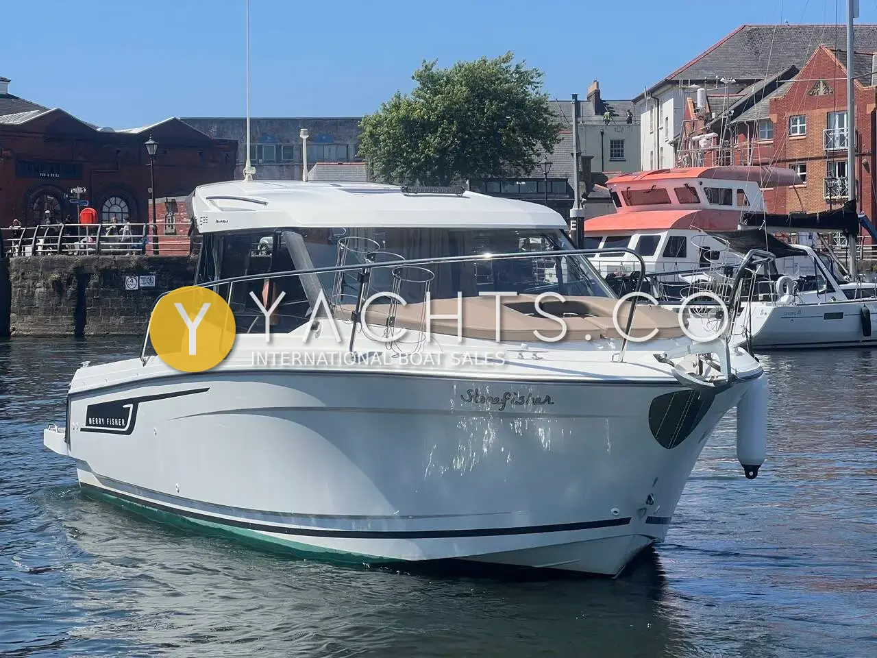 Jeanneau Merry Fisher 695: white wheelhouse cruiser seen from the bow quarter, moored in a harbour lined with brick buildings