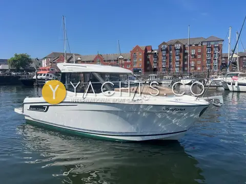 Jeanneau Merry Fisher 695: port bow view of the hull with foredeck sunpad and roof rails, marina apartments behind