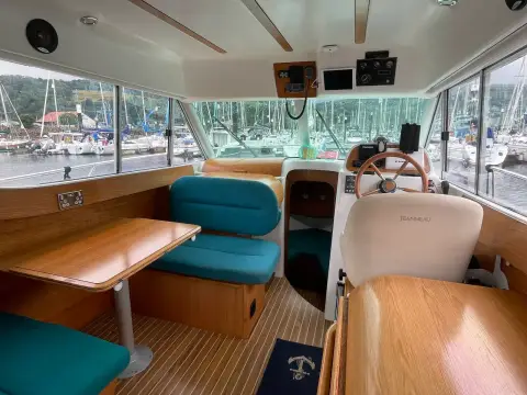 Jeanneau Merry Fisher 805: wheelhouse saloon with blue upholstered dinette, wooden table and helm station facing marina views