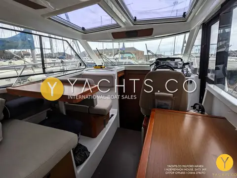 Jeanneau Merry Fisher 855: saloon with wooden dinette table, helm seat and large windows under twin roof hatches