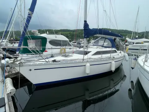 Jeanneau Sun Dream 28: starboard side of the hull afloat in the marina with blue sprayhood and wooded hills behind
