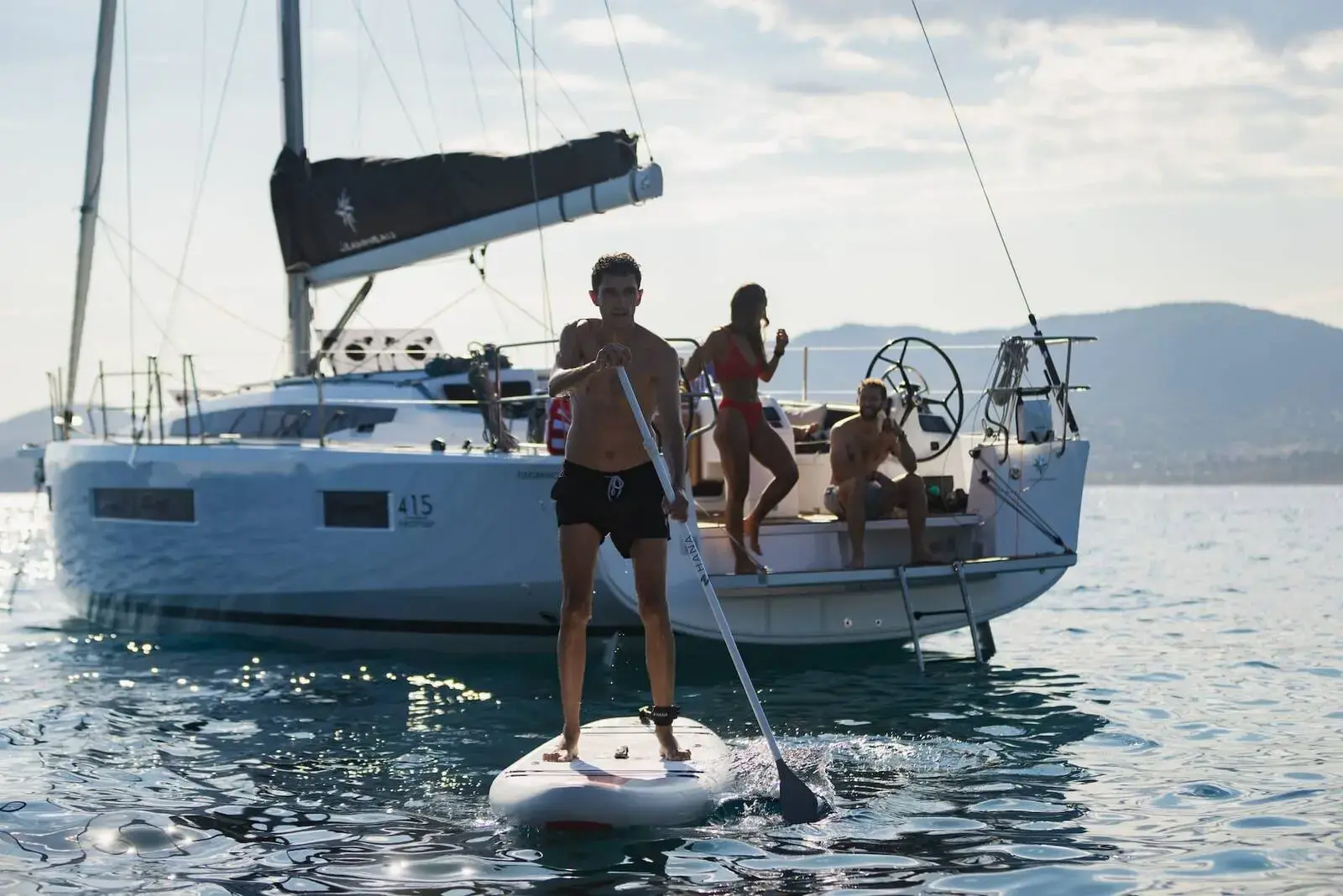 Jeanneau Sun Odyssey 415: white hull at anchor with guests on the transom platform and a paddleboarder alongside