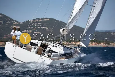 Jeanneau Sun Odyssey 440: heeled under sail from astern, crew at the starboard wheel, wooded hillside behind