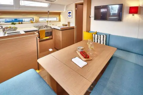 Jeanneau Sun Odyssey 490: saloon with blue upholstered seating, wooden table and galley behind, television on bulkhead