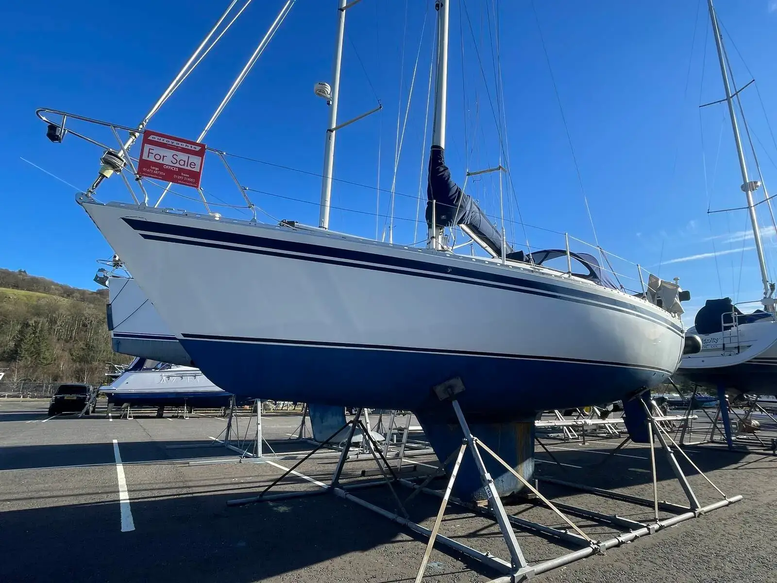Moody 31: hull from the bow quarter, white topsides with blue stripes, ashore on a cradle in a boatyard