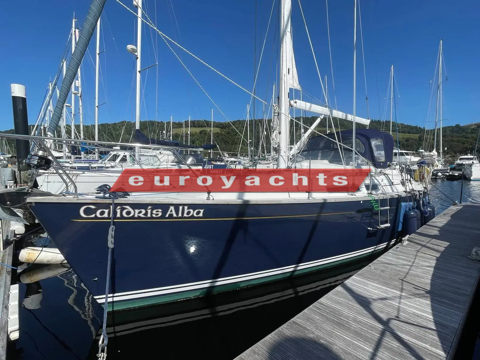 Moody 38 CC Caledonia: dark blue hull with name on topsides, moored stern-to on a pontoon with green hills behind