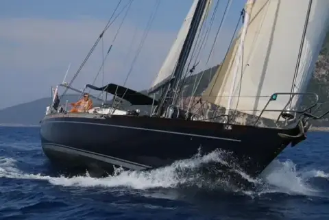 Mystic 60: bow-on view under sail, navy hull with white cove stripe cutting through waves