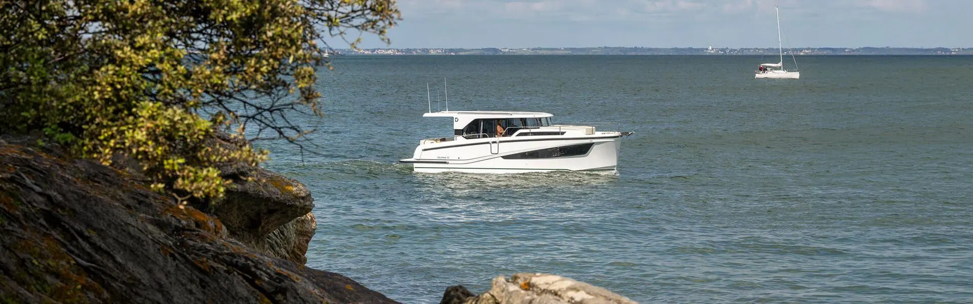 Delphia 10 Sedan: white motor cruiser at anchor in open water, seen past rocks and a tree, sailing yacht beyond