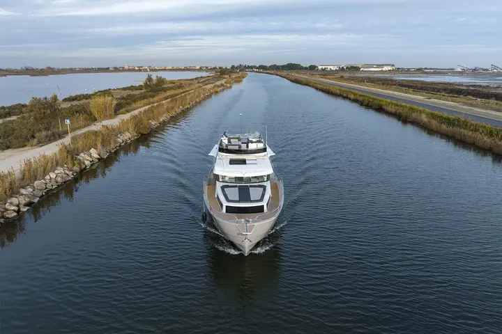 Delphia 12 FlyLounge: aerial bow view of the yacht cruising along a straight canal between grassy banks