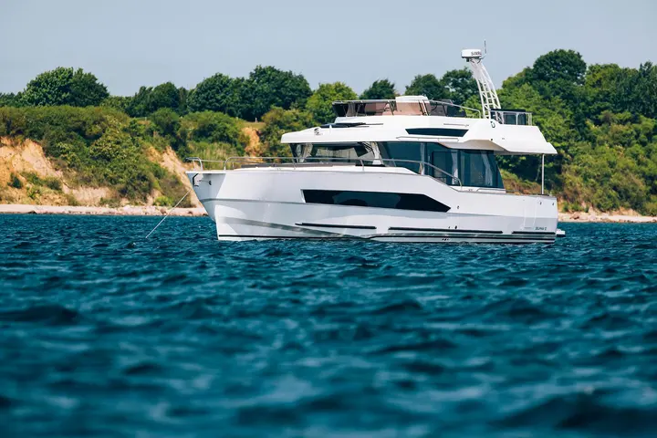 Delphia 12 FlyLounge: white motor yacht with flybridge and radar arch at anchor off a wooded shoreline