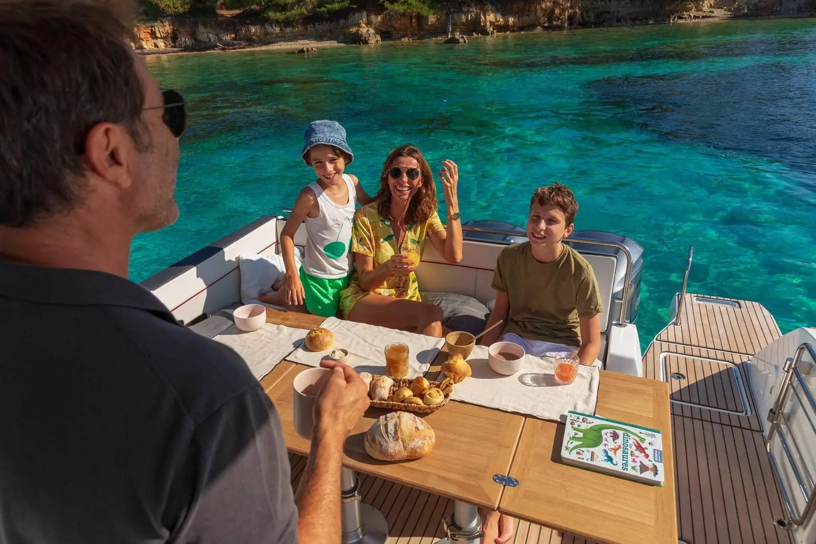 Jeanneau Cap Camarat 10.5 WA Serie 2: cockpit with family seated around a wooden table laid for breakfast, turquoise water beyond the stern