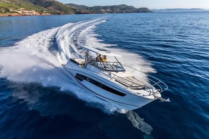 Jeanneau Cap Camarat 10.5 WA Serie 2: underway near a coastline, two people at the helm under the hardtop, foredeck sunpad visible