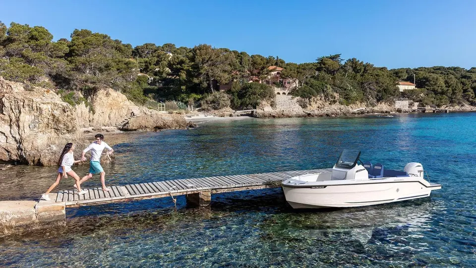 Jeanneau Cap Camarat 6.0 CC: white centre console boat moored beside a wooden jetty in a clear rocky cove, two people running along the jetty