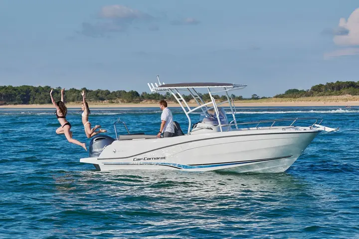 Jeanneau Cap Camarat 6.5 CC: white centre console boat with T-top at sea, two women jumping off the stern near a sandy shore