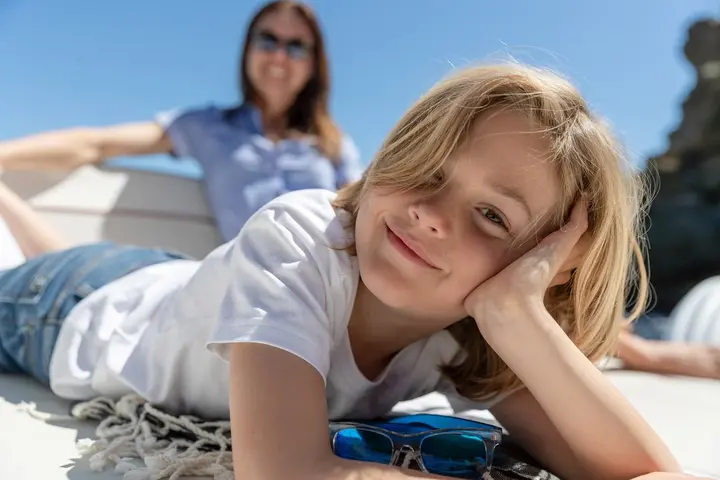 Jeanneau Cap Camarat 6.5 WA: smiling child lying on a towel on the deck with blue sunglasses, woman relaxing behind