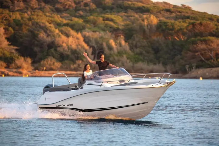 Jeanneau Cap Camarat 6.5 WA: white motorboat with black hull stripes cruising past a wooded shore, two people at the helm