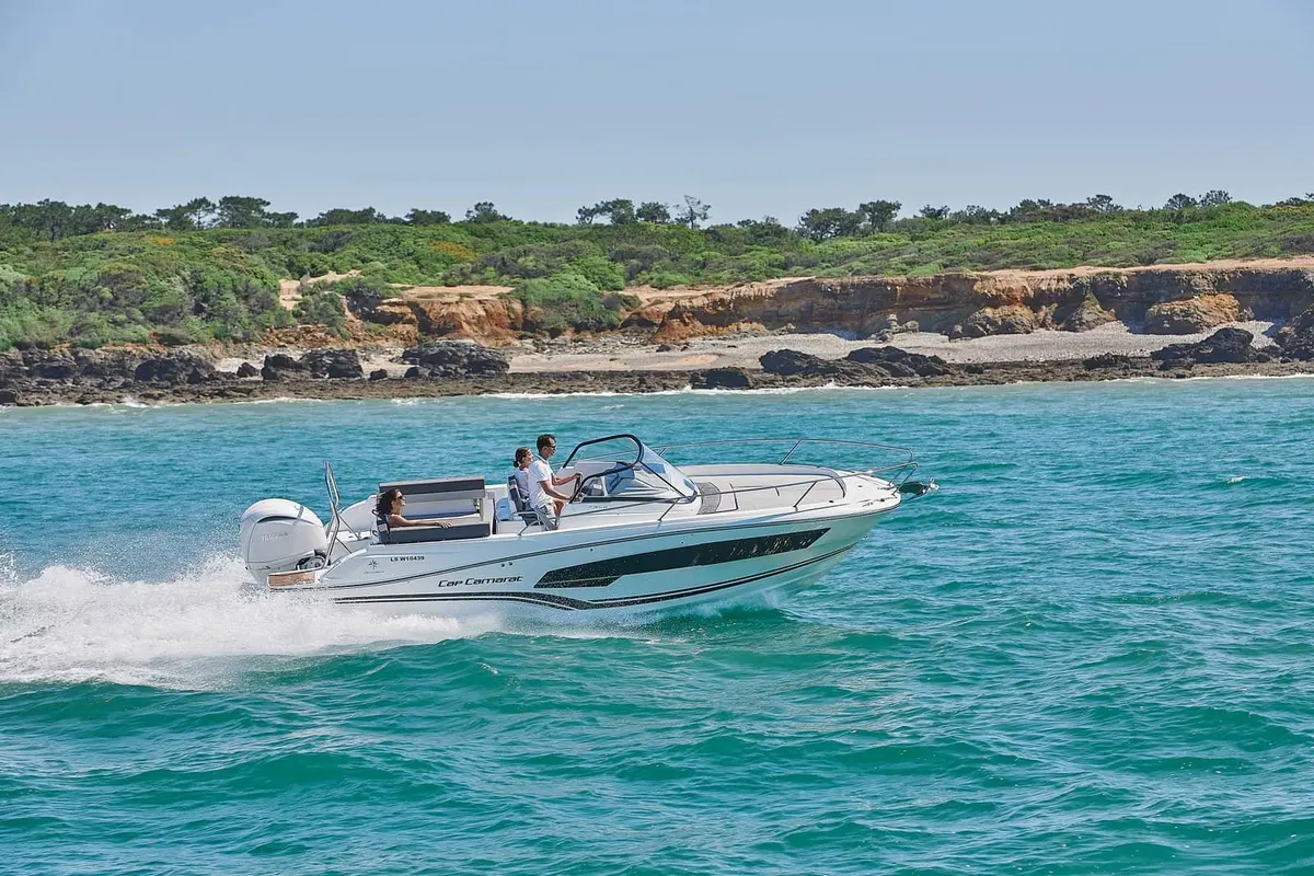 Jeanneau Cap Camarat 7.5 WA: white motorboat under way at speed with three people aboard, rocky coastline behind
