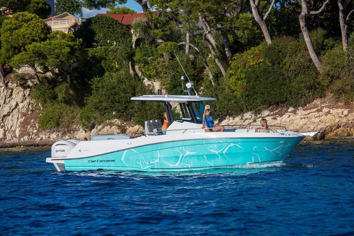 Jeanneau Cap Camarat 9.0 CC Serie 2: centre console boat with turquoise patterned hull and hardtop cruising past a rocky wooded shoreline
