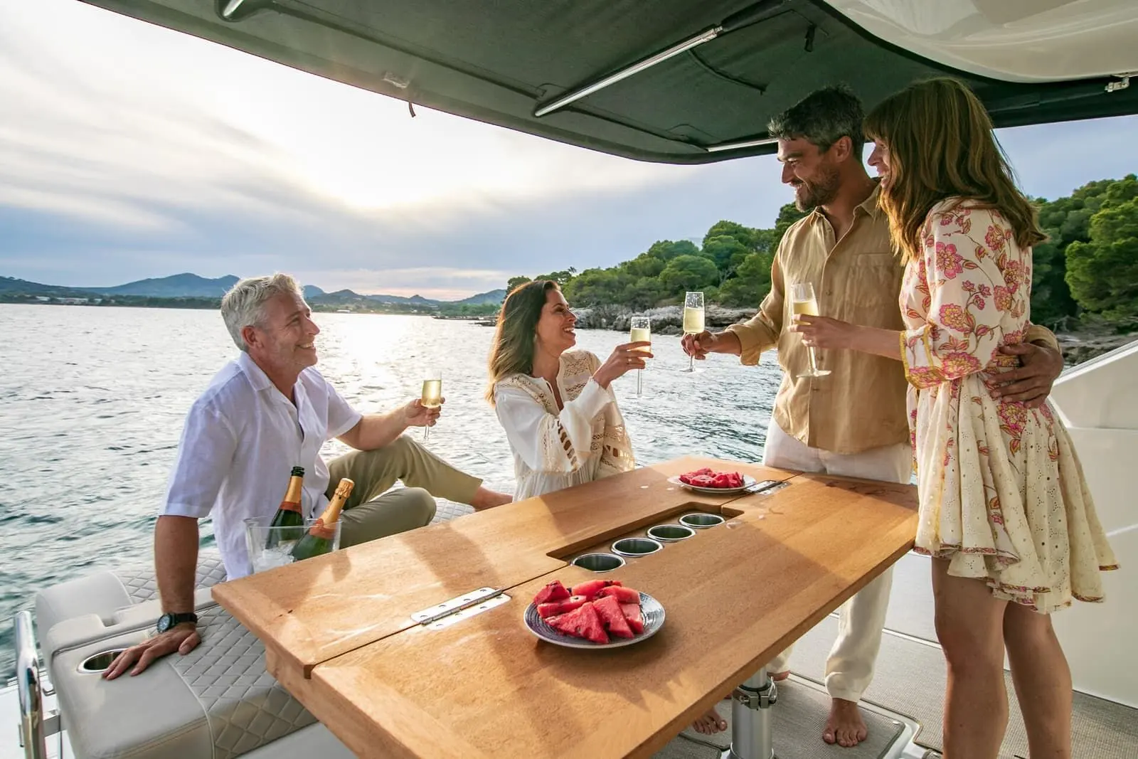 Jeanneau DB 37 IB: four people toasting with glasses around the wooden cockpit table under the hardtop, watermelon on plates