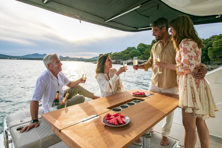 Jeanneau DB 37 IB: four people toasting with glasses around the wooden cockpit table under the hardtop, watermelon on plates