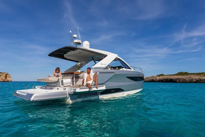 Jeanneau DB 37 IB: stern view at anchor in turquoise water, man seated on fold-out side terrace, woman on aft sunpad