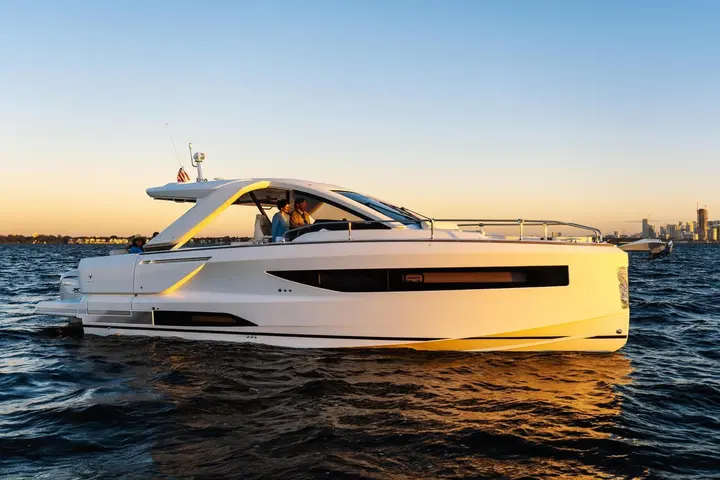 Jeanneau DB 37 OB: side profile underway at sunset, people at helm under hardtop, city skyline in background