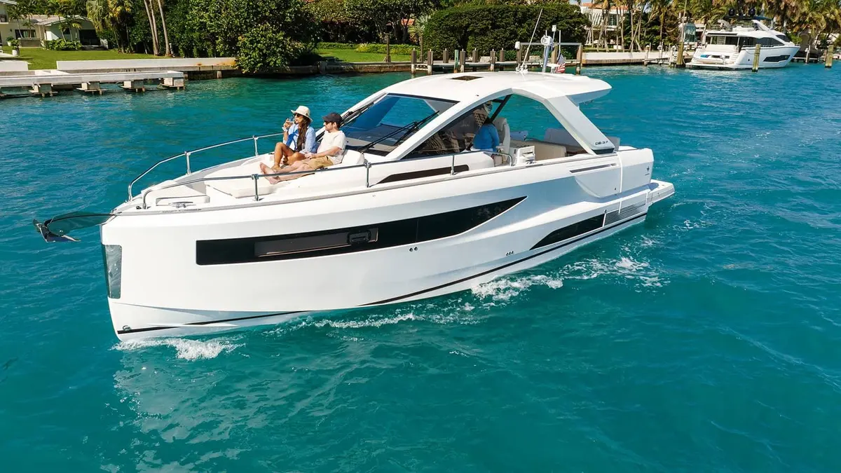 Jeanneau DB 37 OB: white motor yacht cruising on turquoise water, two people relaxing on foredeck sunpad near hardtop