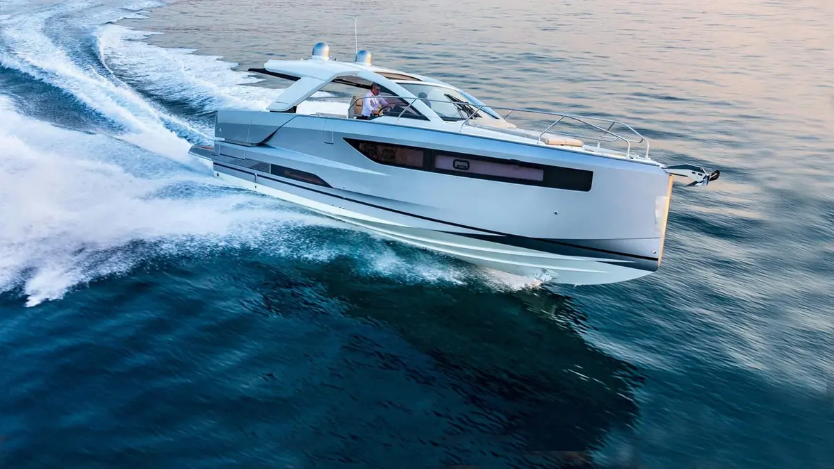 Jeanneau DB 43 IB: silver motor cruiser under way at speed, skipper at the helm under a hardtop, white wake behind