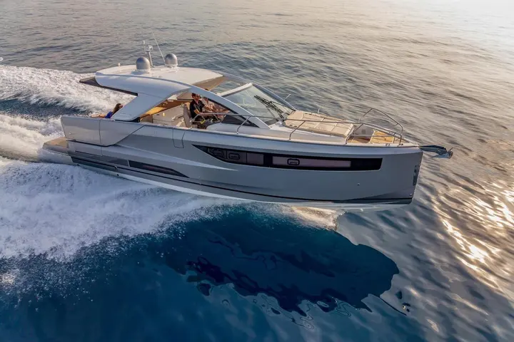 Jeanneau DB 43 IB: silver motor yacht under way from above, skipper at the helm, passenger aft, sunpads on foredeck