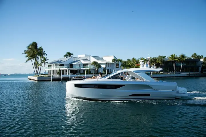 Jeanneau DB 43 OB: white hull cruising past waterfront villa and palm trees, passengers seated at helm and foredeck