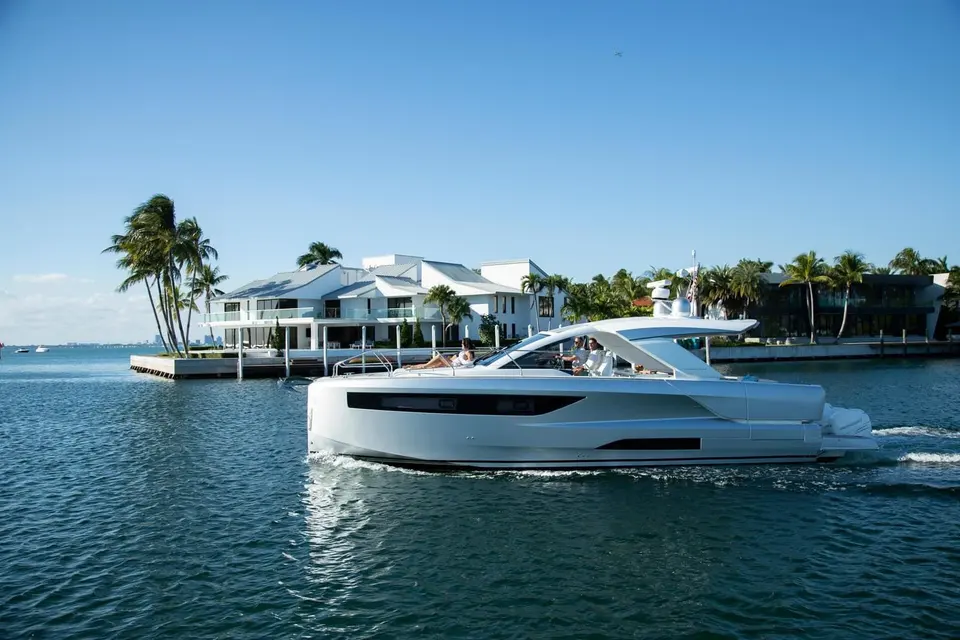 Jeanneau DB 43 OB: white hull cruising past waterfront villa and palm trees, passengers seated at helm and foredeck