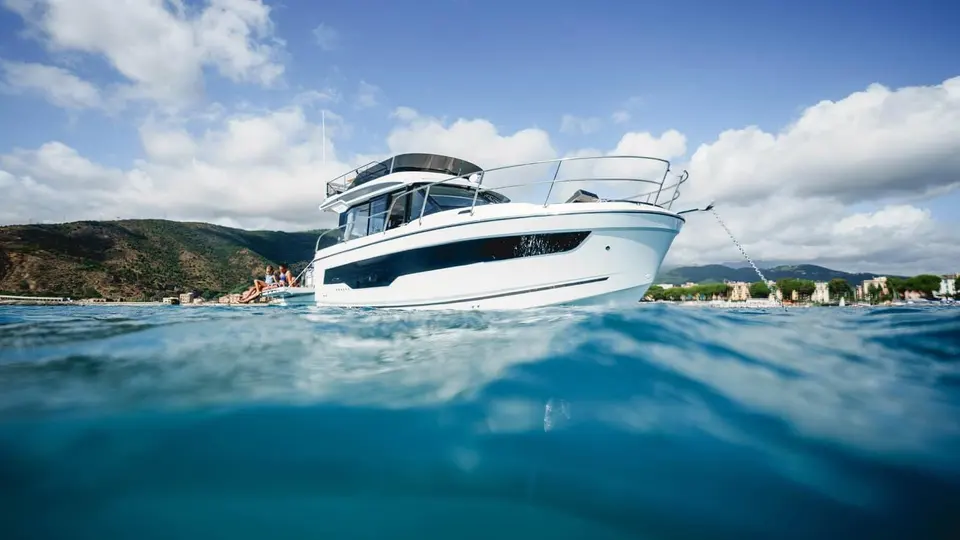 Jeanneau Merry Fisher 1095 Fly SERIE2: starboard side at anchor near a hilly coastline, showing flybridge, hull windows and bow anchor chain