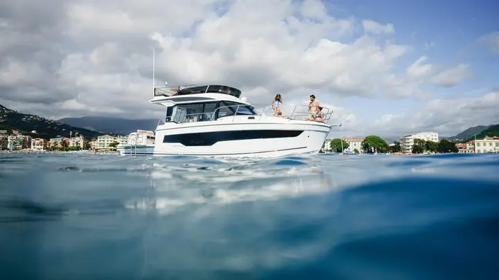 Jeanneau Merry Fisher 1095 Fly SERIE2: white flybridge motor boat at anchor near a coastal town, three people standing on the foredeck