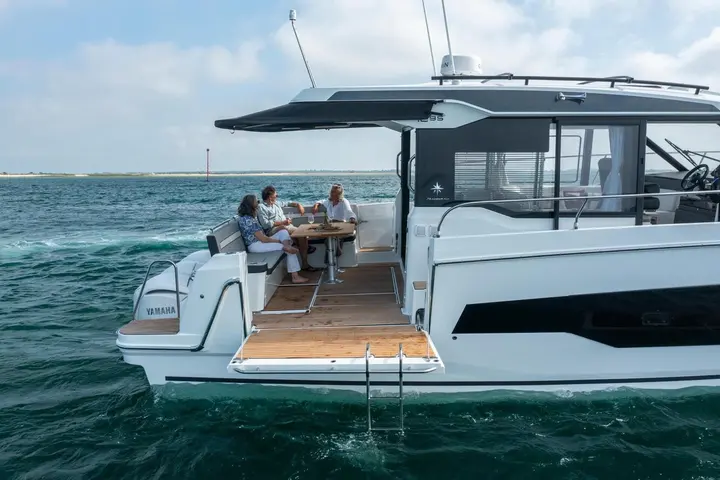 Jeanneau Merry Fisher 1295 Coupe: aft cockpit with three people seated at a teak table, wooden decking and bathing platform ladder